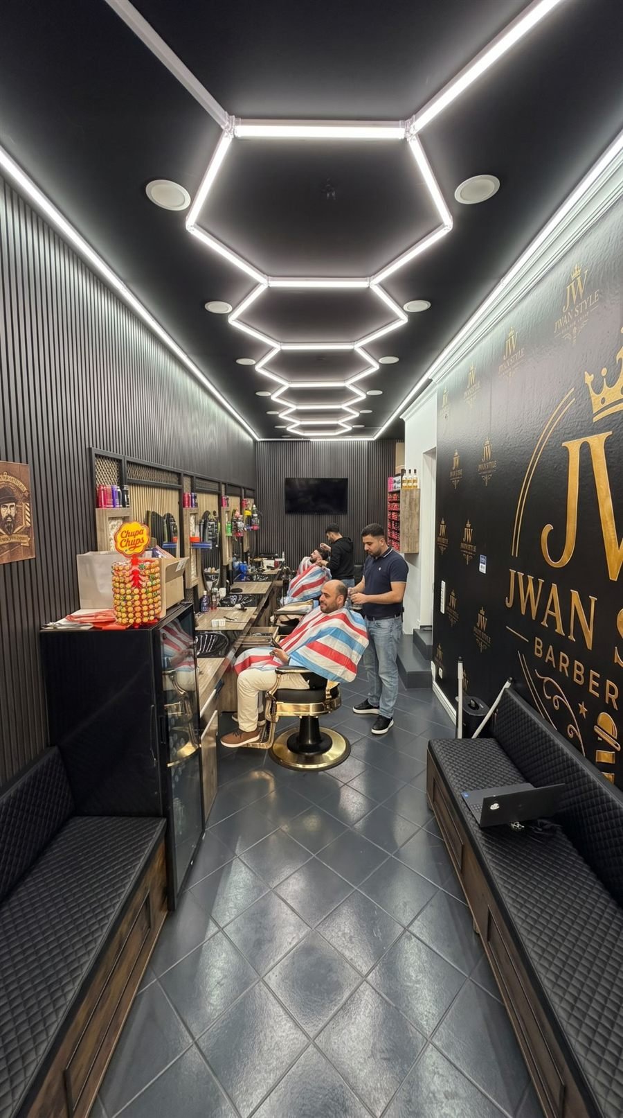 Innenansicht JWAN STYLE Barber Shop in Karlsruhe: Barberstühle, Wabenlicht an der Decke und Logo-Wand in Gold
